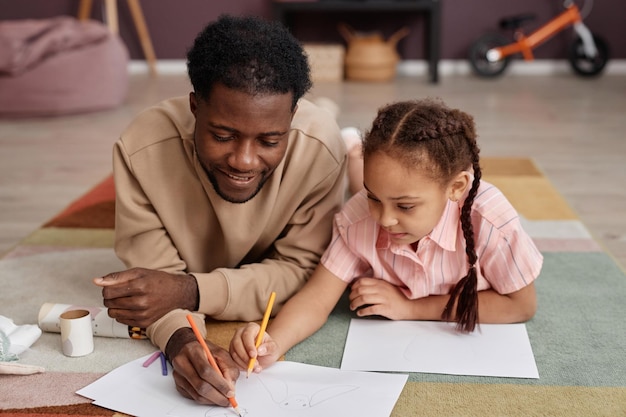 Pai e filha negros desenhando fotos deitados no chão em casa e sorrindo