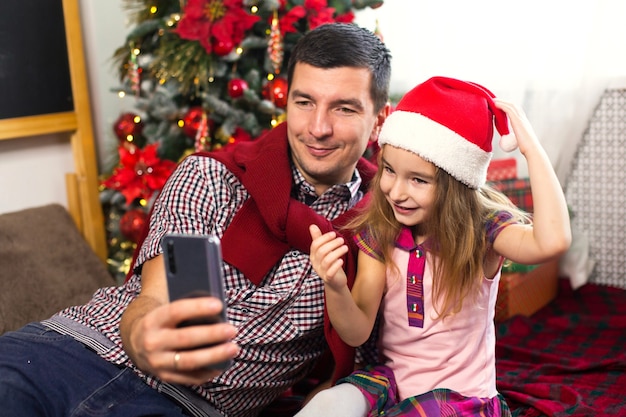 Pai e filha perto da árvore de natal com um smartphone fazem uma selfie e se comunicam via conexão de vídeo. saudações de natal, uma caixa de presente, uma garota com um chapéu de papai noel acenando olá. ano novo, decoração de feriado