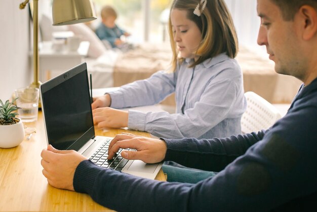 Pai e filha sentados à mesa em casa. Homem de negócios, trabalhar no laptop, garota escreve em um caderno. Conceito de educação online, trabalho remoto