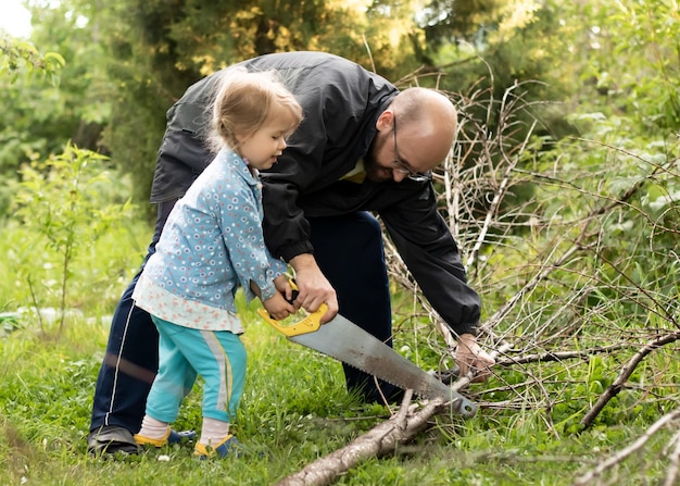 Pai e filha trabalham juntos no jardim, cortando galhos de árvores secas com serra manual
