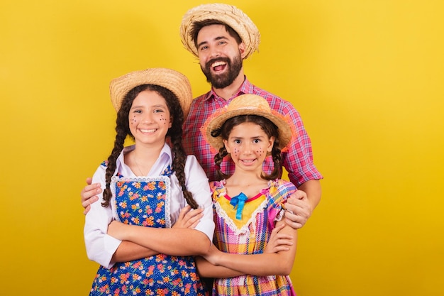 Pai e filhas vestindo roupas típicas da Festa Junina abraçados sorrindo olhando para a câmera Para a festa do Arraia