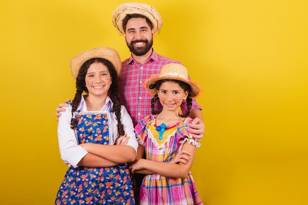 Pai e filhas vestindo roupas típicas da Festa Junina abraçados sorrindo olhando para a câmera Para a festa do Arraia