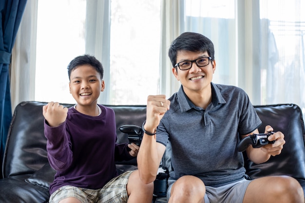 Pai e filho asiáticos gostam de jogar videogame com joystick de vídeo com emoção e muita diversão na sala de estar em casa