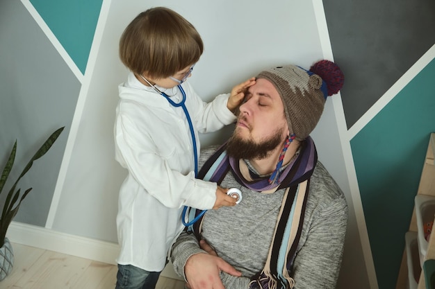 Pai e filho brincando de clínica e médico menino de vestido médico com estetoscópio trata o pai