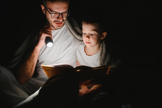 Pai e filho com lanterna lendo livro debaixo do cobertor em casa