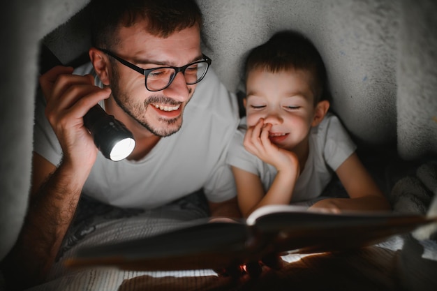 Pai e filho com lanterna lendo livro debaixo do cobertor em casa