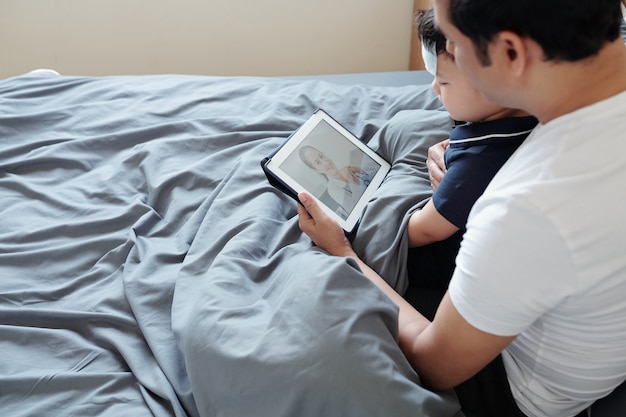 Pai e filho doente sentados na cama com tablet digital e pediatra com videochamada para pedir conselhos