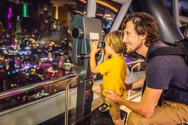 Pai e filho estão olhando para a paisagem urbana de Kuala Lumpur Use binóculos Vista panorâmica do horizonte da cidade de Kuala Lumpur à noite ao pôr do sol na construção de arranha-céus na Malásia Viajando com o conceito de crianças