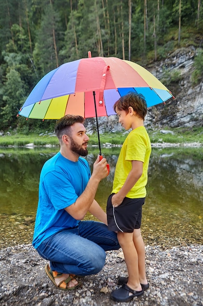 Pai e filho estão passando o dia dos pais na zona rural perto do rio, pai está segurando um guarda-chuva acima de seu filho para protegê-lo da chuva.