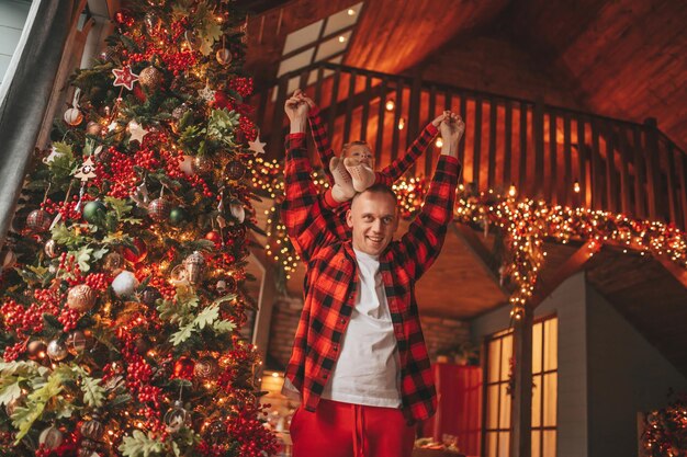 Pai e filho felizes autênticos e sinceros em pijama xadrez vermelho brincando no alojamento de madeira Natal decorado