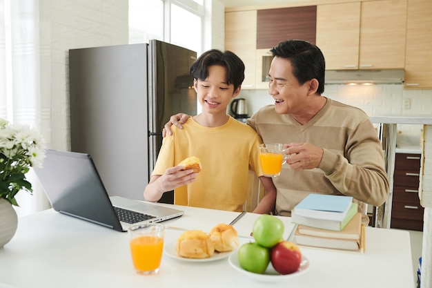 Pai e filho felizes desfrutando de pães frescos com suco de laranja e discutindo um bom dia na escola