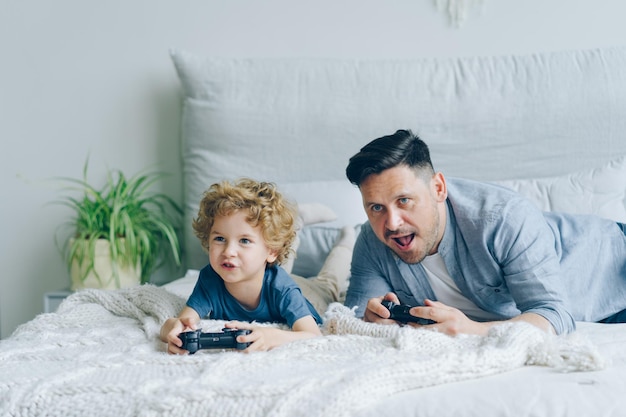 Pai e filho jogando videogame na cama em casa pressionando botões no joystick