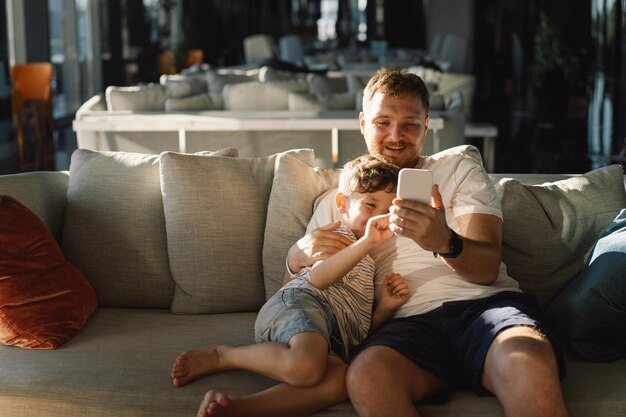 Pai e filho sentam-se relaxando no sofá no corredor do hotel divertindo-se usando o telemóvel