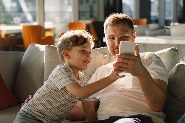 Pai e filho sentam-se relaxando no sofá no corredor do hotel divertindo-se usando o telemóvel