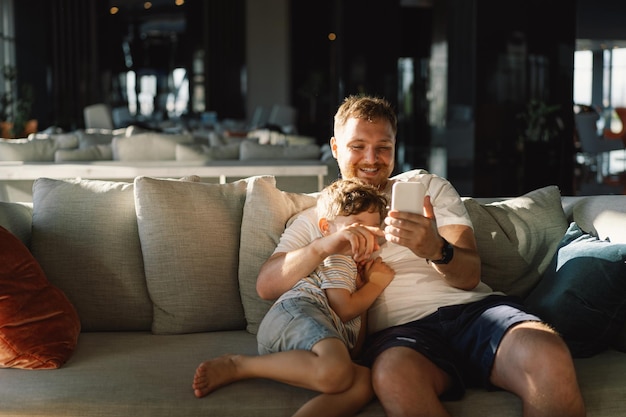 Pai e filho sentam-se relaxando no sofá no corredor do hotel divertindo-se usando o telemóvel