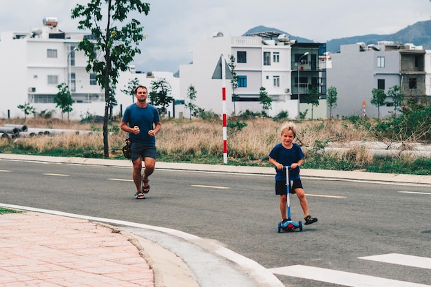 Foto pai e filho vão a desportos de manhã. o homem corre, a criança anda de scooter. aldeia moderna.