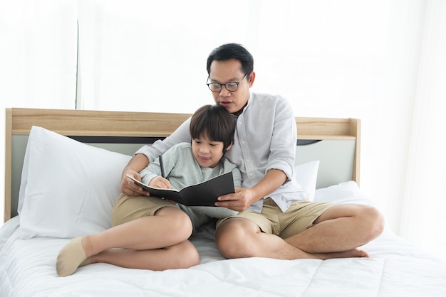 Pai e menino asiático escrevendo no livro, deitado na cama em casa, pai e filho passando bons momentos juntos.