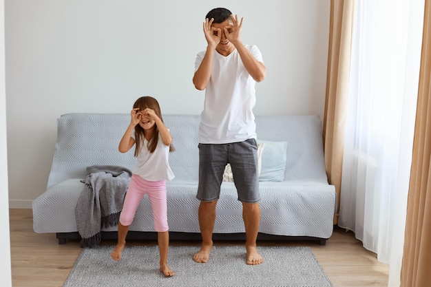 Pai engraçado positivo vestindo jeans curto e camiseta branca, dançando com sua filha contra o sofá na sala de estar, fazendo binóculos com as mãos, olhando para a câmera.