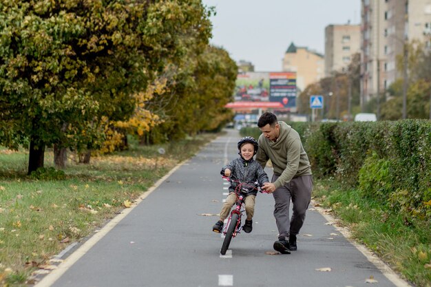 Pai ensina o filho a andar de bicicleta na ciclovia do parque. O pai está segurando uma bicicleta e o filho está sentado nela.