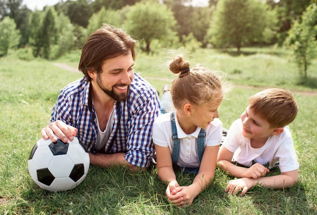 Pai está deitado na grama no Prado com crianças. Ele está segurando uma bola com a mão. Homem está olhando para seu filho com sua filha. Rapaz parece feliz e engraçado.