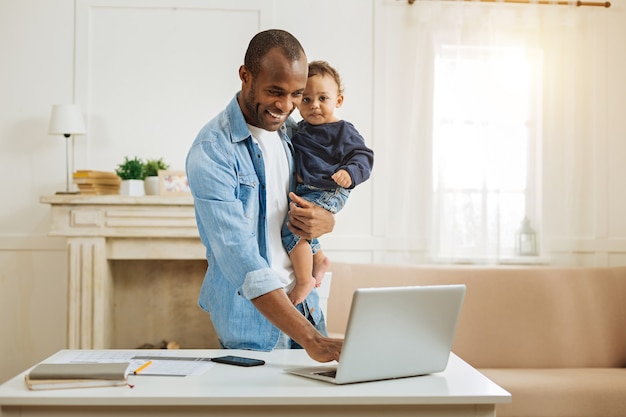 Pai feliz. bonito e inspirado jovem pai afro-americano segurando seu filho pequeno e digitando no laptop enquanto está à mesa com uma lareira ao fundo