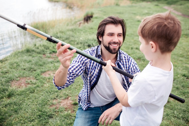 Pai feliz está dando vara de peixe para seu filho. ele está sorrindo. rapaz está segurando a vara de peixe muito apertada e olhando para o pai.