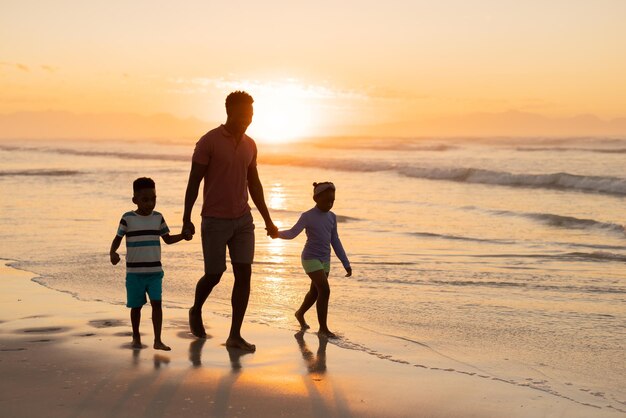Pai jovem afro-americano segurando as mãos do filho e da filha enquanto caminhava na praia contra o céu