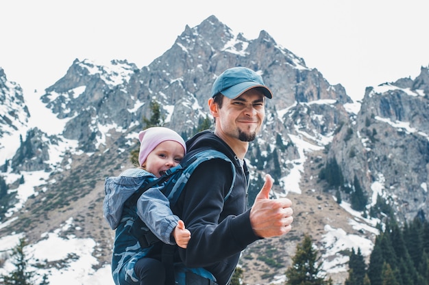 Pai jovem e atraente com sua filha bebê em um marsúpio ergonômico nas costas viaja pela montanha