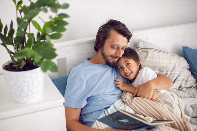 Pai lê um livro deitado em uma cama branca para seu filho antes de ir para a cama