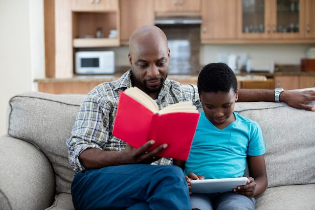 Pai lendo livro enquanto filho sentado ao lado dele usando tablet digital