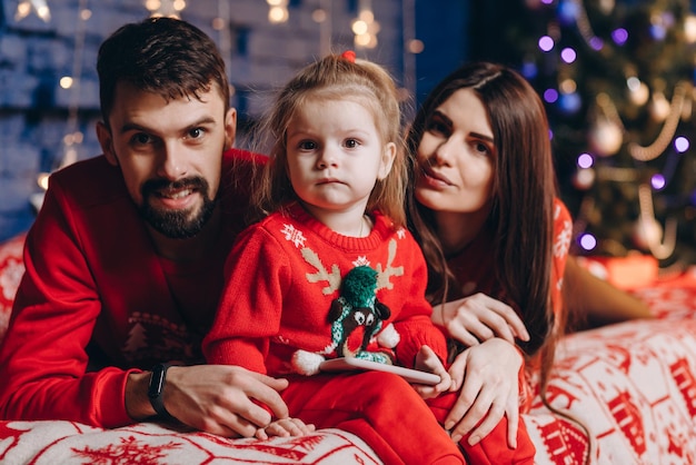 Pai mãe e filha estão deitados na cama em frente à árvore de natal