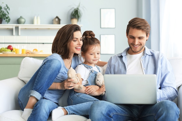 Foto pai, mãe e filha usando dispositivos eletrônicos, sentados no sofá da sala.