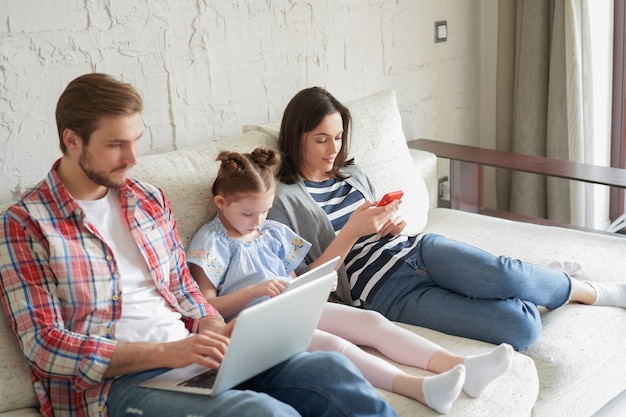 Pai, mãe e filha usando dispositivos eletrônicos, sentados no sofá da sala.