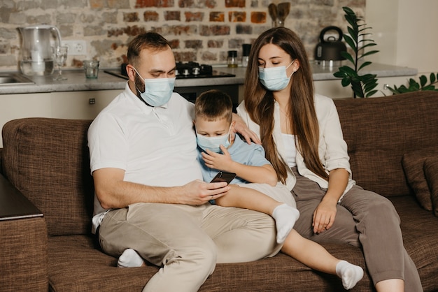 Pai, mãe e filho estão sentados em um sofá com máscaras médicas para evitar a propagação do coronavírus (COVID-19). Família durante a quarentena em casa. Papai está dando notícias para seus parentes