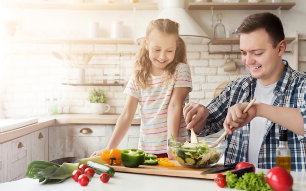 Pai positivo e filha cozinhando na cozinha em casa. homem ensinando menina a preparar uma salada saudável, copie o espaço