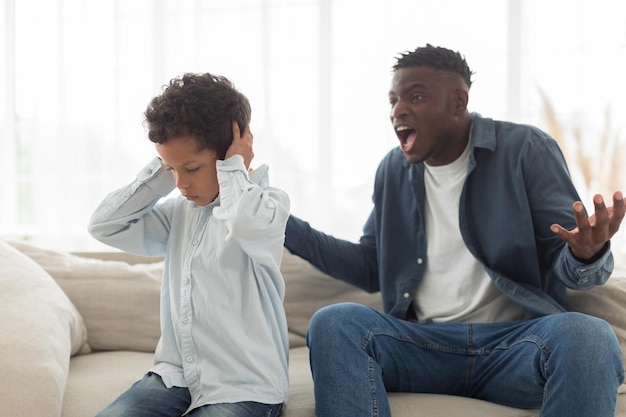 Pai preto zangado a gritar com o filho assustado em casa.