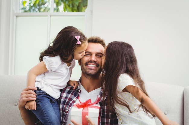 Pai segurando uma caixa de presente e suas filhas beijando-o