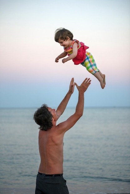 Pai sem camisa carregando filha enquanto está de pé na praia contra o céu