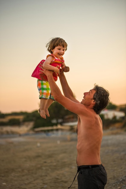 Pai sem camisa carregando filha na praia durante o pôr do sol