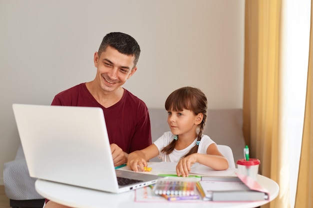 Pai sorridente e uma filha fazendo lição de casa em casa, sentados à mesa na sala, olhando para a tela do laptop com sorrisos, tendo emoções positivas, educação online.