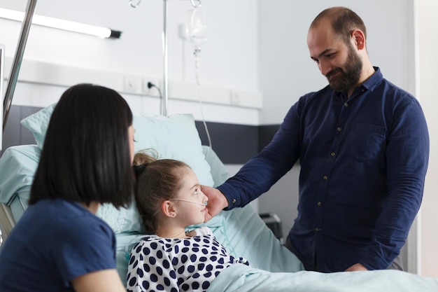 Pai sorridente feliz tocando o rosto da filha jovem doente. Adorável jovem pai olhando para a menina doente e tocando seu rosto enquanto está sentado na sala da enfermaria de recuperação clínica pediátrica.