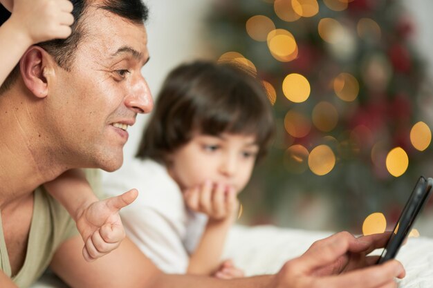 Foto pai sorridente usando tablet digital enquanto passa um tempo com seus filhinhos fofos, deitado na cama em casa decorada para o natal. tecnologia, conceito de paternidade