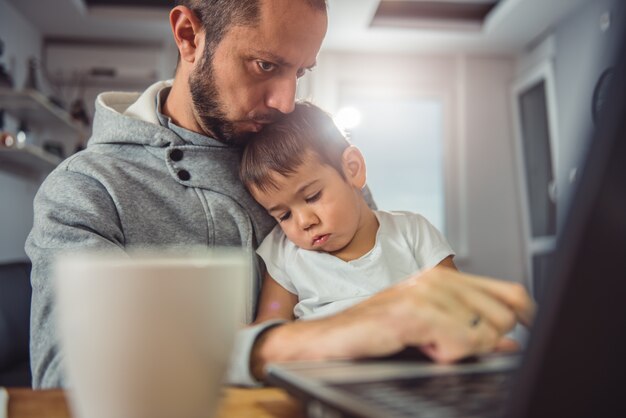 Pai trabalhando no laptop e segurando o filho no colo