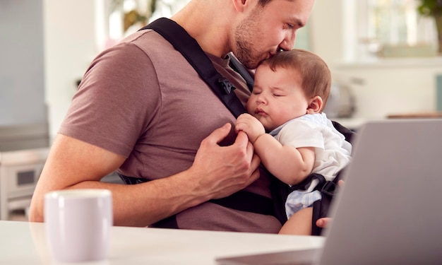 Pai transgênero trabalhando em casa no laptop cuidando do filho bebê no estilingue