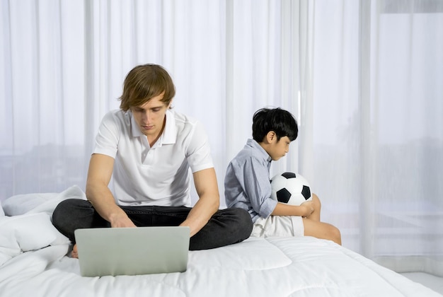 Pai usando computador portátil para trabalhar no quarto enquanto seu filho espera de lado com uma bola de futebol