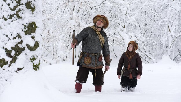 Pai viking com filho na floresta de inverno Pai conversando com filho segurando machado nas mãos