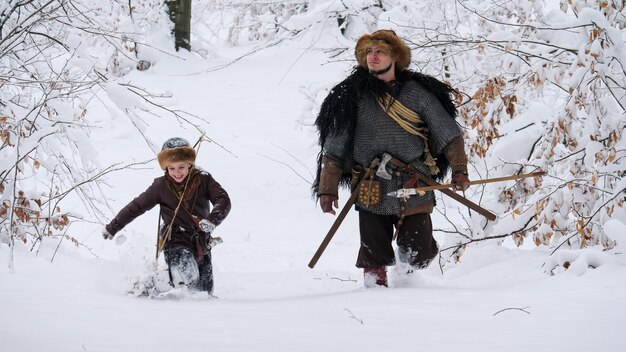 Pai viking com seu filho indo na floresta de inverno, eles tendo machado, lança, cebola. Eles se vestiram com roupas medievais.