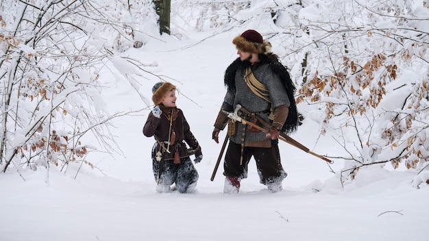 Pai viking com seu filho indo na floresta de inverno, eles tendo machado, lança, cebola.