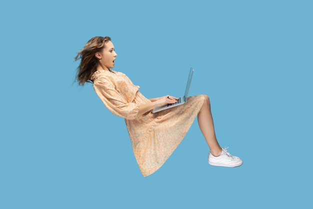 Pairando no ar Garota surpreendida no vestido amarelo levitando olhando para a tela do laptop chocou espantado surfando nas redes sociais da web enquanto voava no ar em estúdio interior isolado em fundo azul