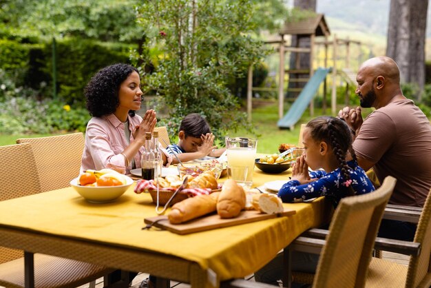 Pais bi-raciais felizes, filho e filha a rezar antes da refeição, sentados à mesa num jardim ensolarado. Verão, família, solidariedade, religião, comida e estilo de vida, inalterados.